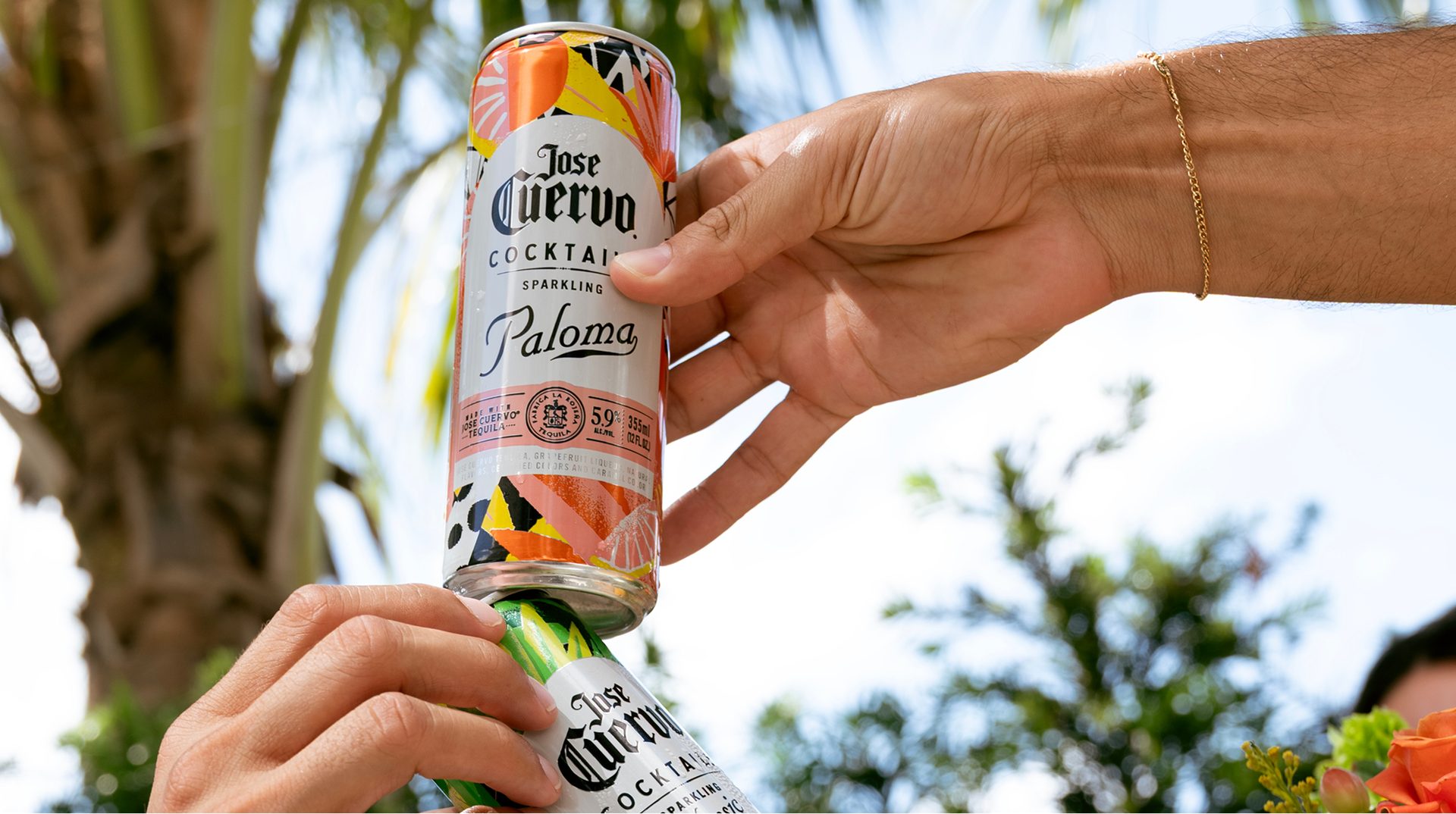 A hand passing a Jose Cuervo Sparkling Paloma can to another hand outdoors under palm trees