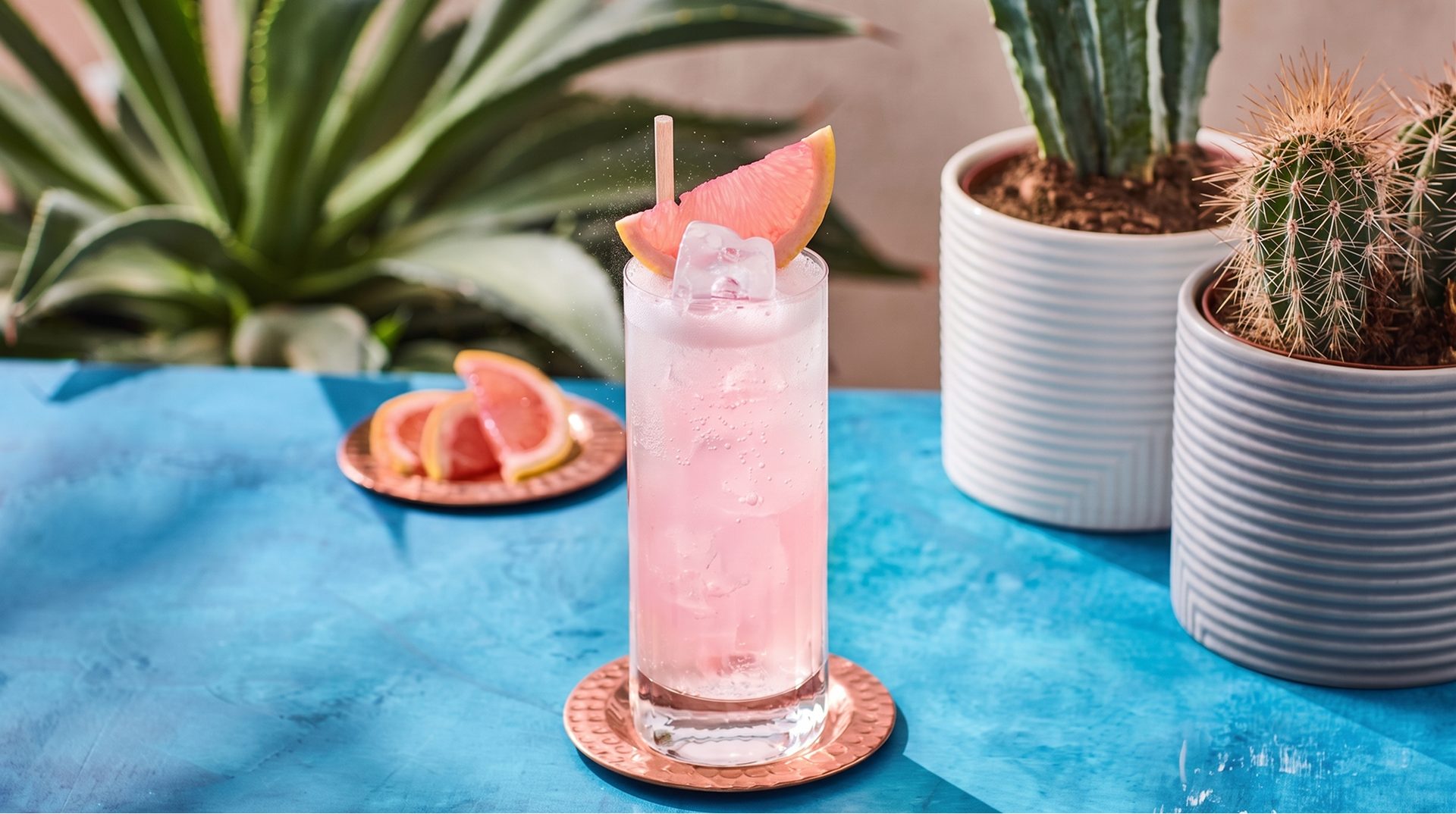 A classic Paloma served over ice in a tall glass with a grapefruit wedge, beside agave and cactus plants