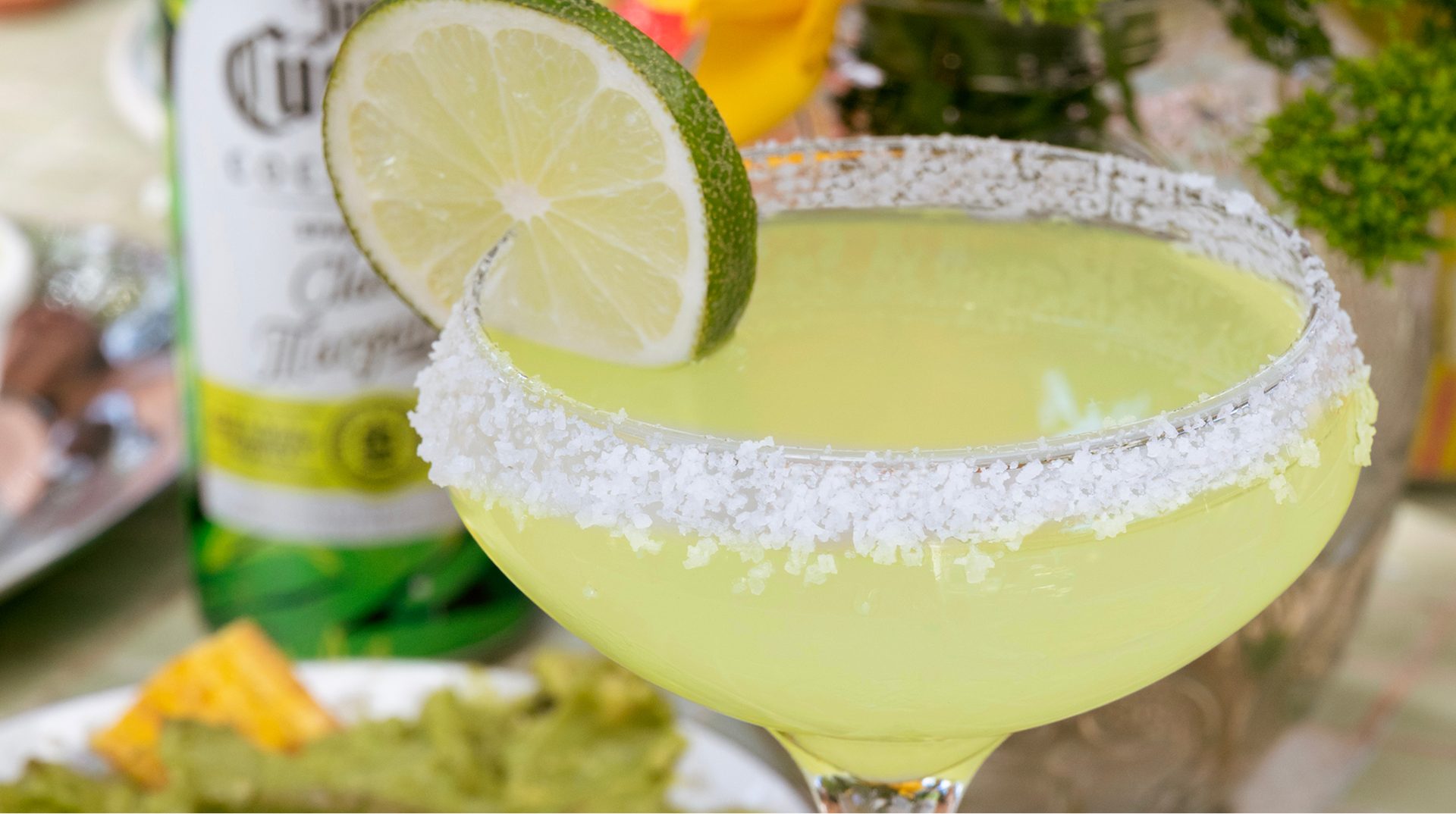 A salt-rimmed margarita garnished with a lime wheel, beside a bottle of Jose Cuervo Classic Margarita
